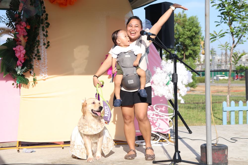 A host on stage with a baby in a carrier and a golden retriever in a cream dress